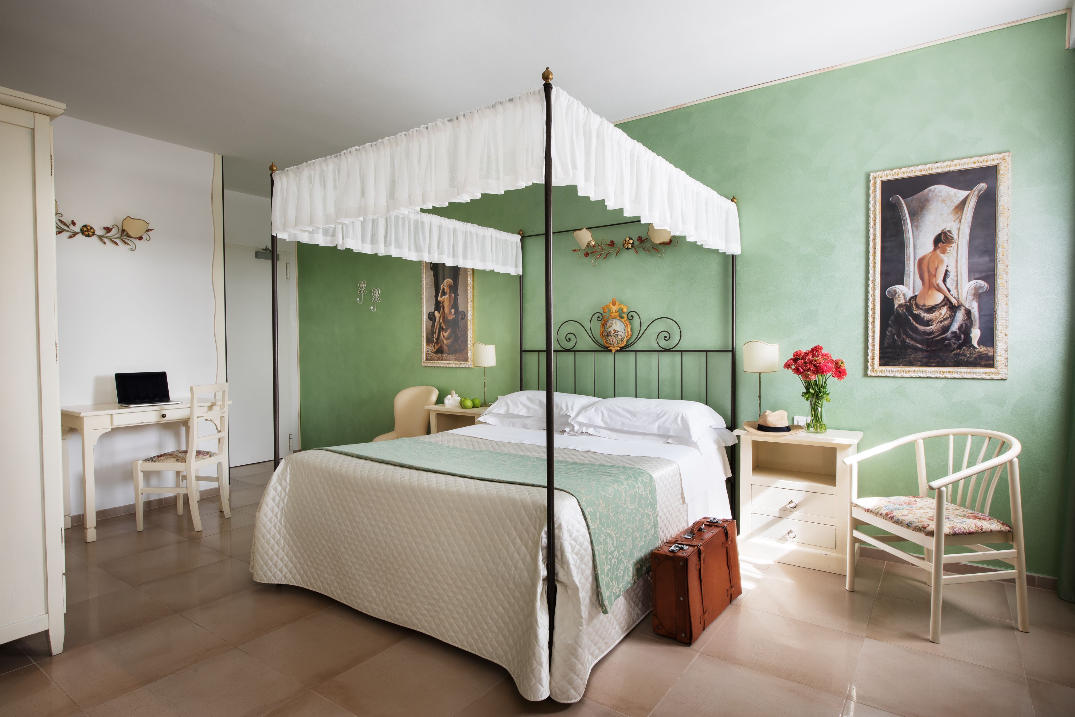 A bedroom with a four-poster bed, a desk, a chair, and a nightstand with a lamp and flowers.