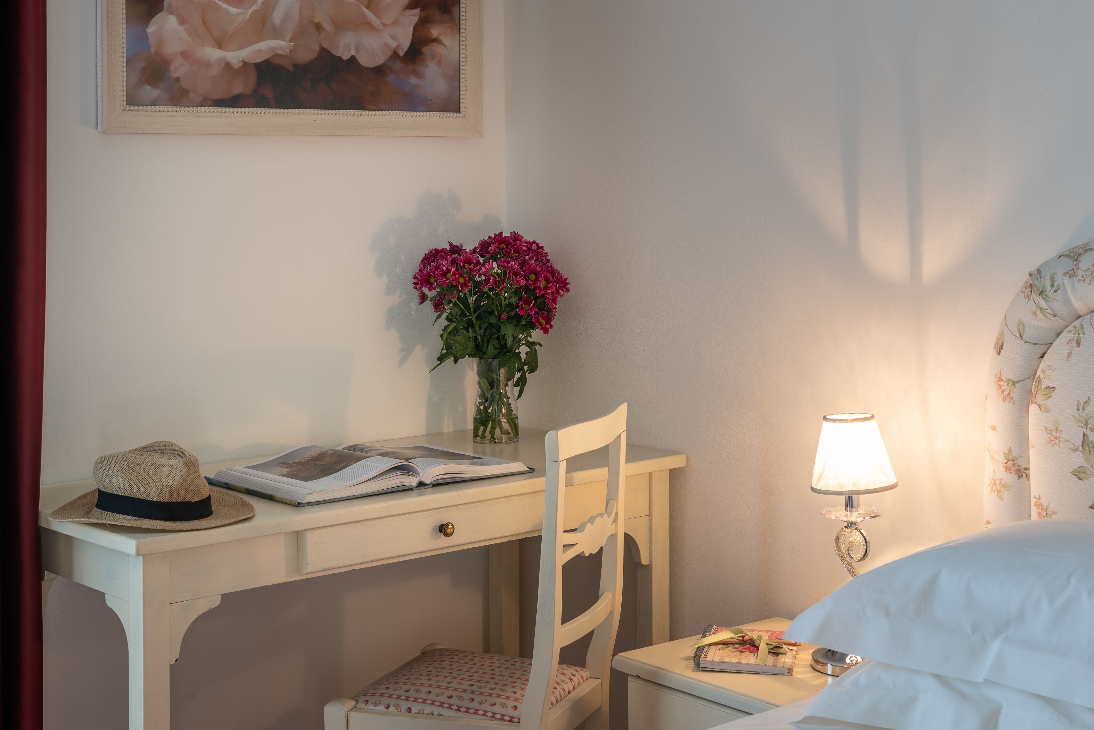 A neatly arranged bedroom with a bed, a bedside table with a lamp, a chair, a desk with a hat and book, and a vase with flowers.
