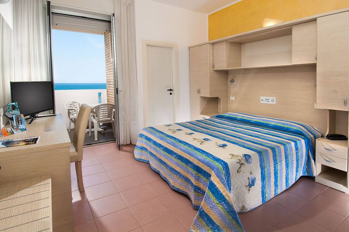 A hotel room with a bed, a desk with a computer, a chair, and a view of the sea.