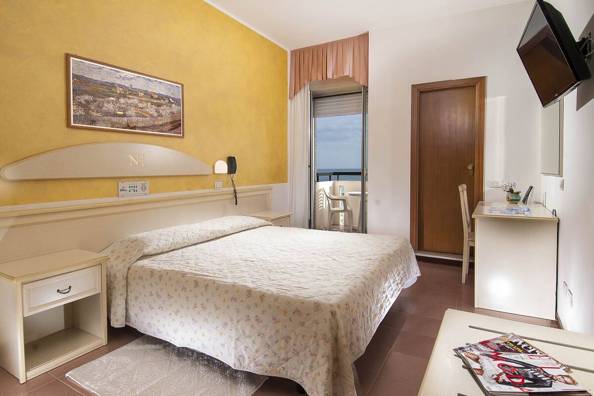 A hotel room with a bed, bedside table, a desk with a chair, a TV, and a balcony with a view.