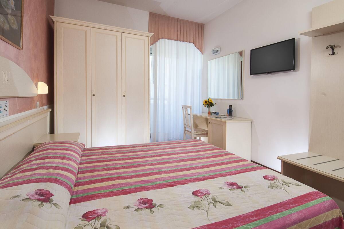 A bedroom with a bed, a desk, a chair, a wardrobe, a television, and a mirror.