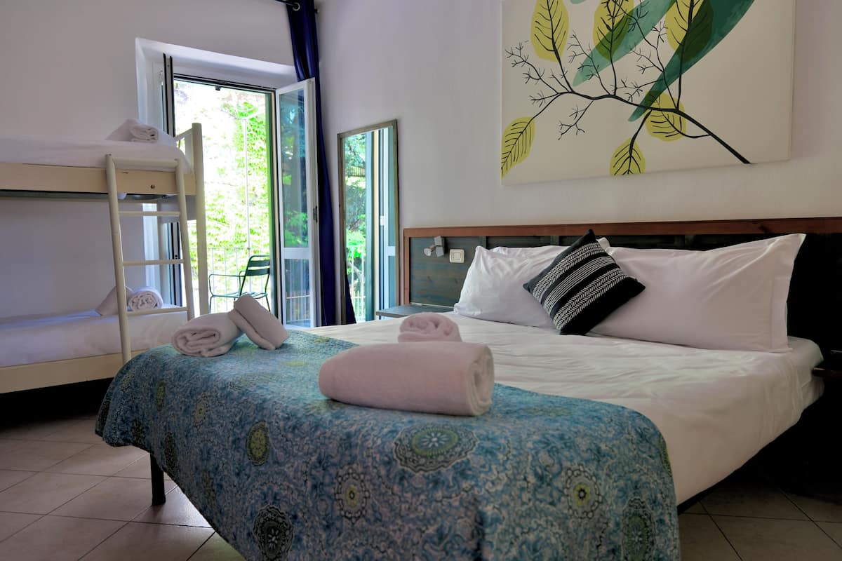 A hotel room with a bunk bed, a single bed with a patterned bedspread, a window with curtains, and a wall-mounted artwork.
