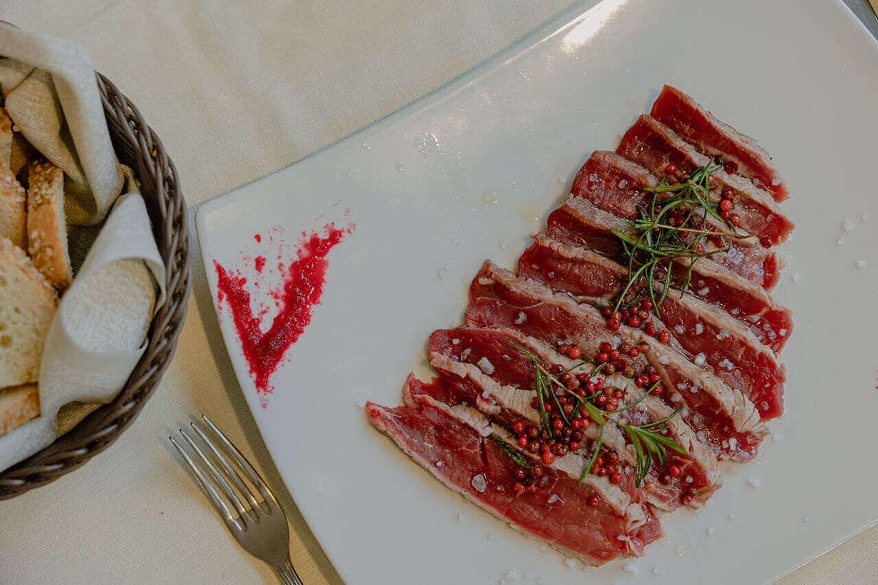 A plate of thinly sliced meat garnished with herbs and peppercorns, accompanied by a basket of bread.