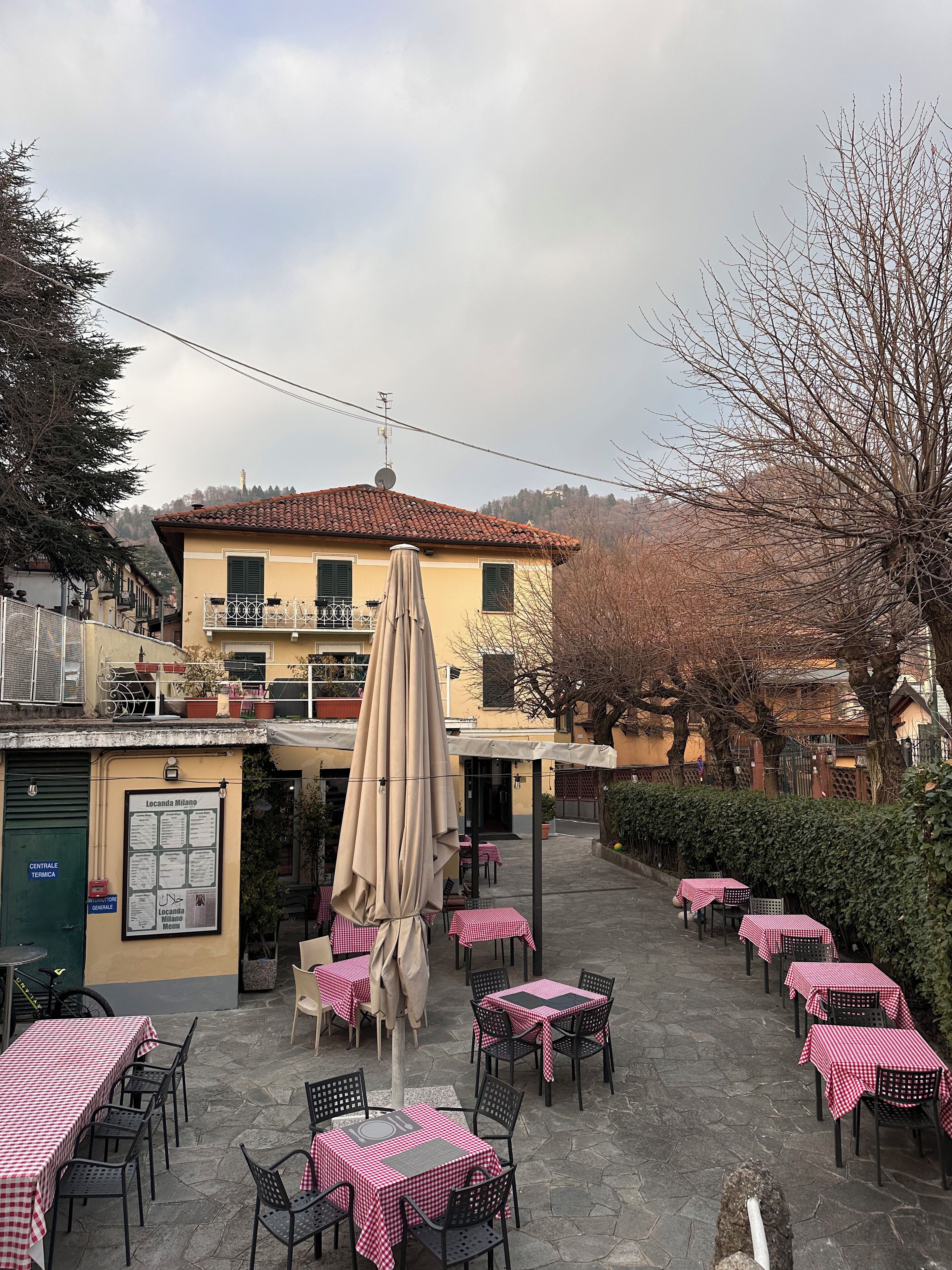 Un café al aire libre con mesas y sillas, un edificio amarillo con techo rojo, y una colina al fondo.