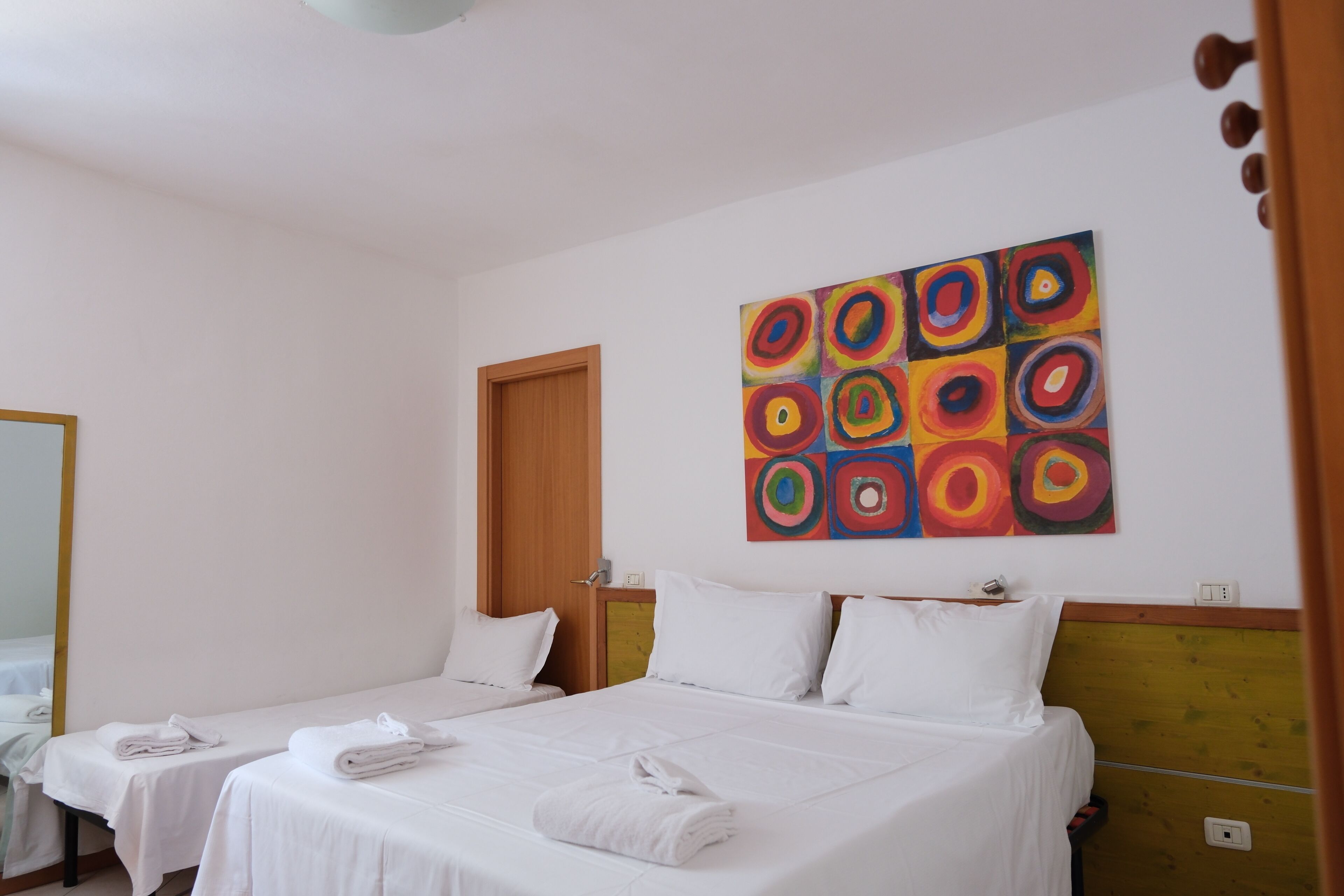A hotel room with two beds, a wooden door, a colorful abstract painting, and a mirror.