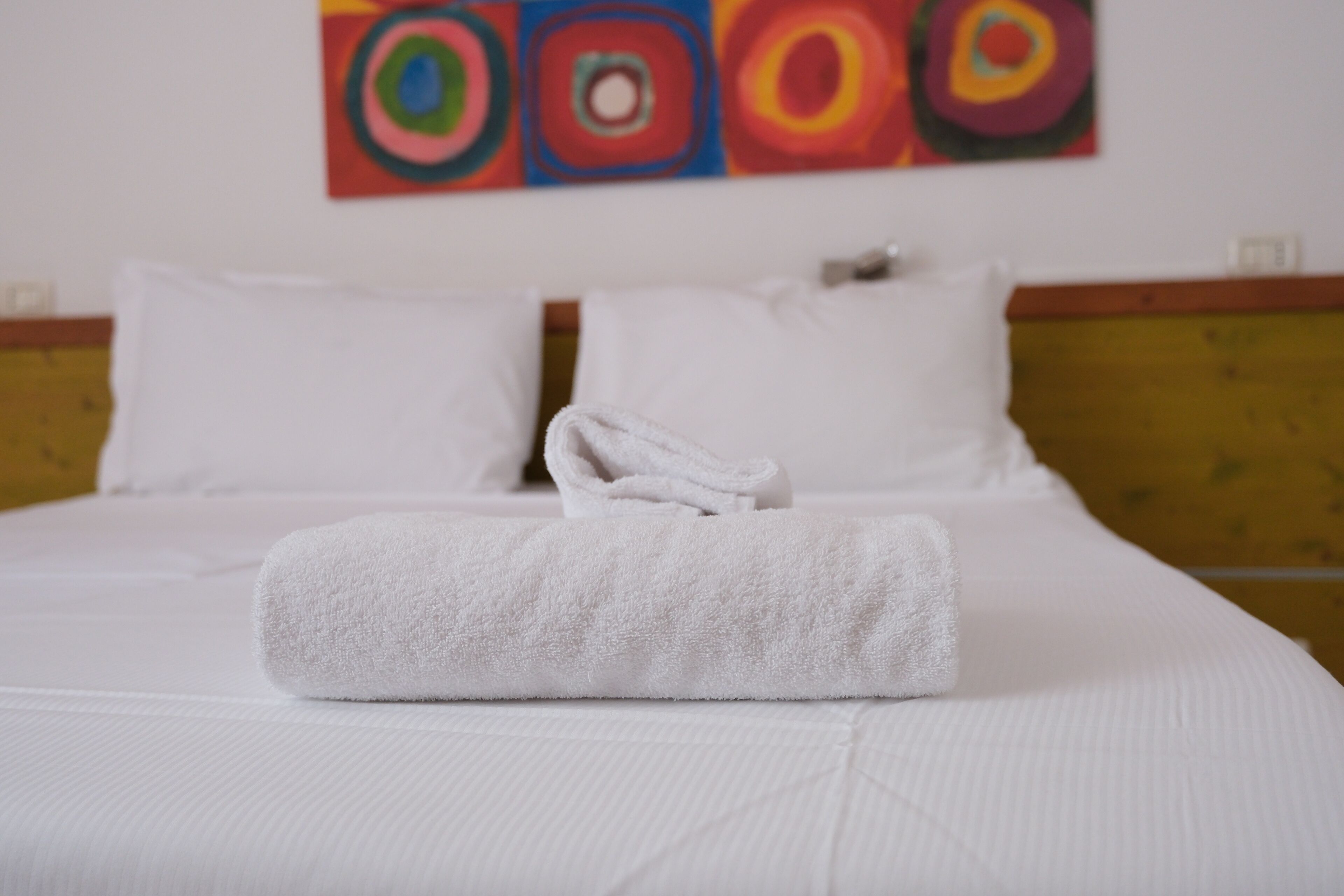 A neatly made bed with white linens and pillows, a rolled white towel, and a colorful abstract painting on the wall.