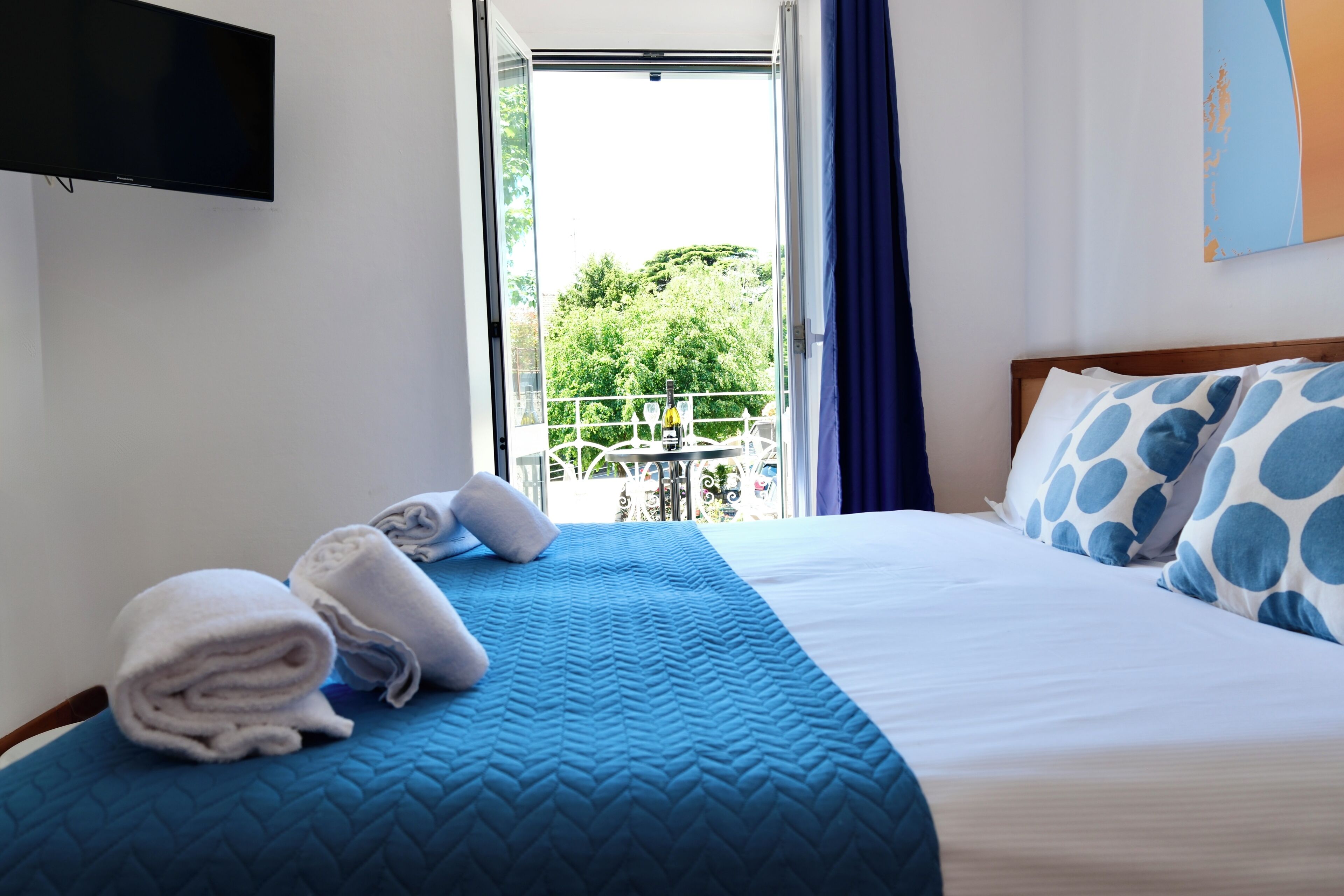 Habitación de hotel con cama, televisor montado en la pared, balcón con barandilla y vista a la vegetación.