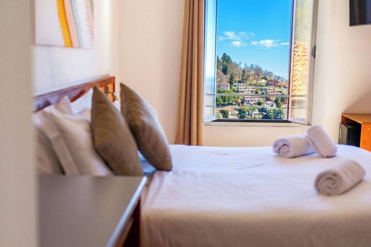 A hotel room with a bed, pillows, a window with a view of a town, and a bedside table.
