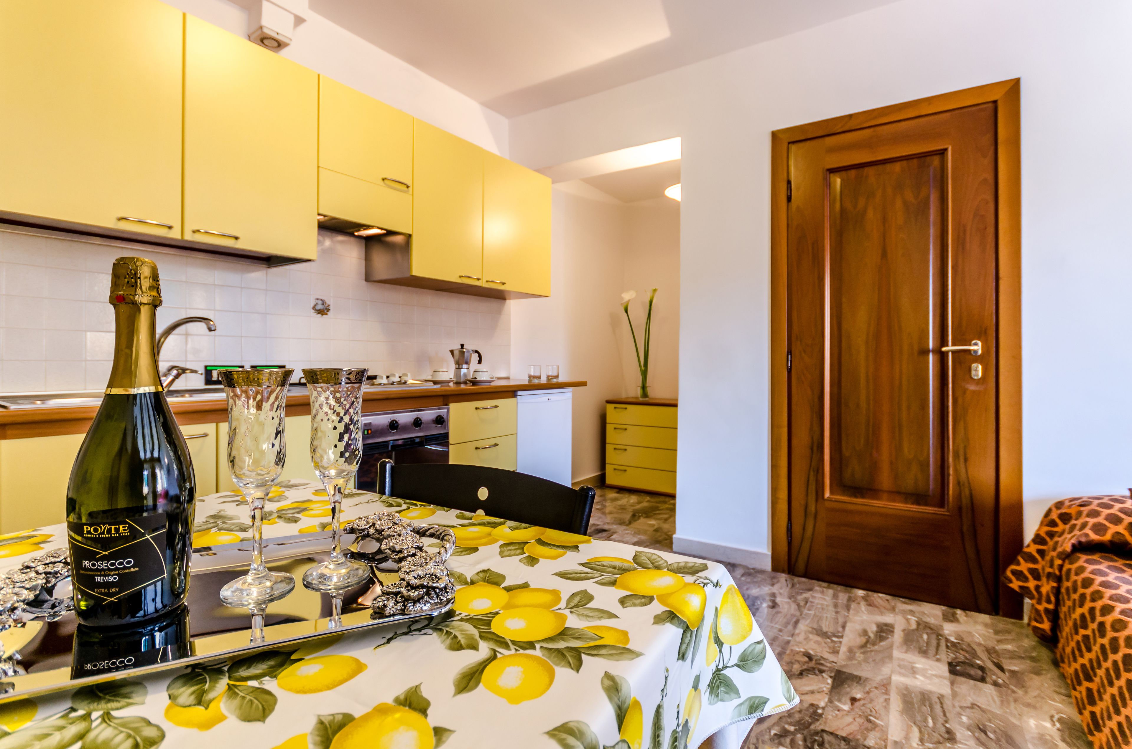 A small kitchen with yellow cabinets, a wooden door, and a dining table with a lemon tablecloth.