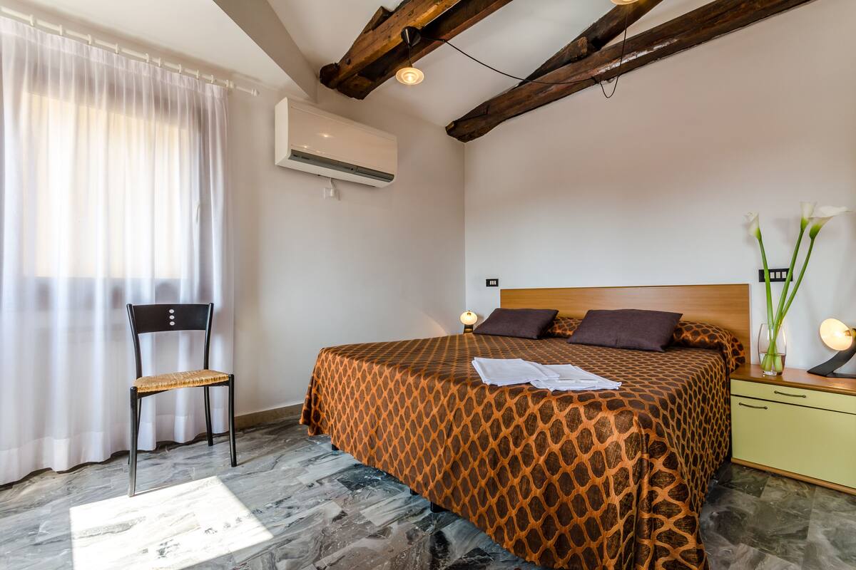 A bedroom with a patterned bedspread, a wooden headboard, a nightstand with a lamp, and a chair.