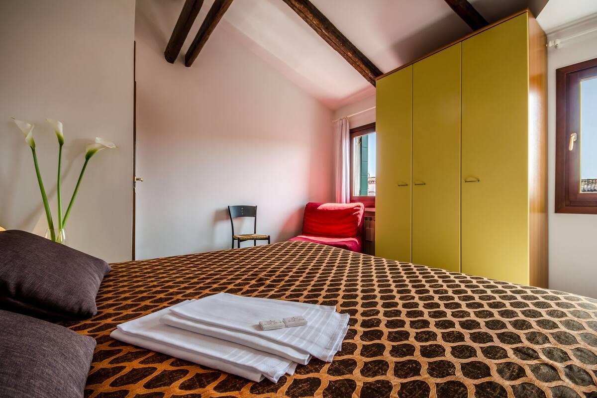 A hotel room with a patterned bedspread, a neatly folded white towel, and a vase with white flowers.