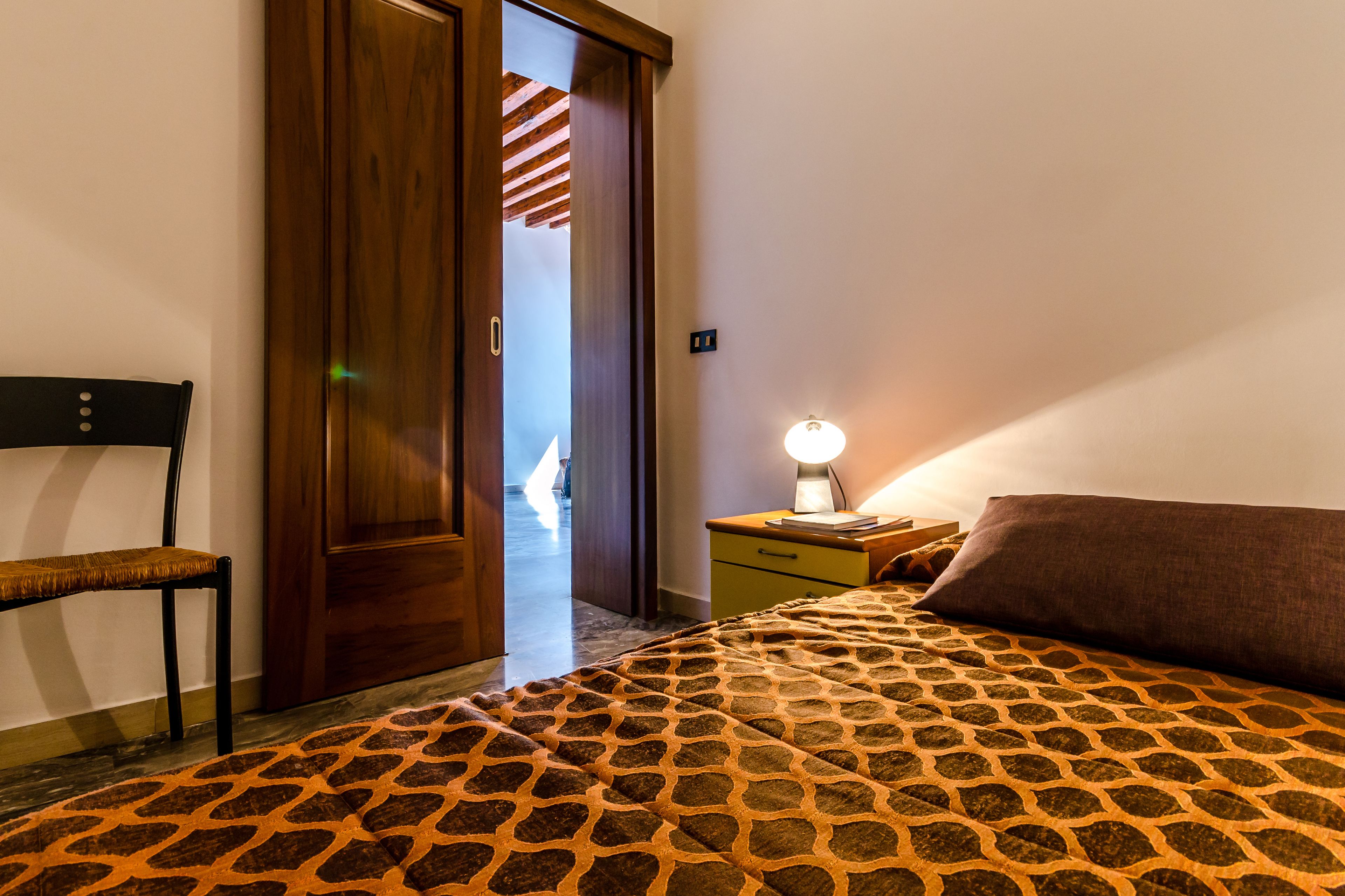 Un dormitorio con una colcha estampada, una silla de madera, una mesita de noche con una lámpara y una puerta que da a otra habitación.