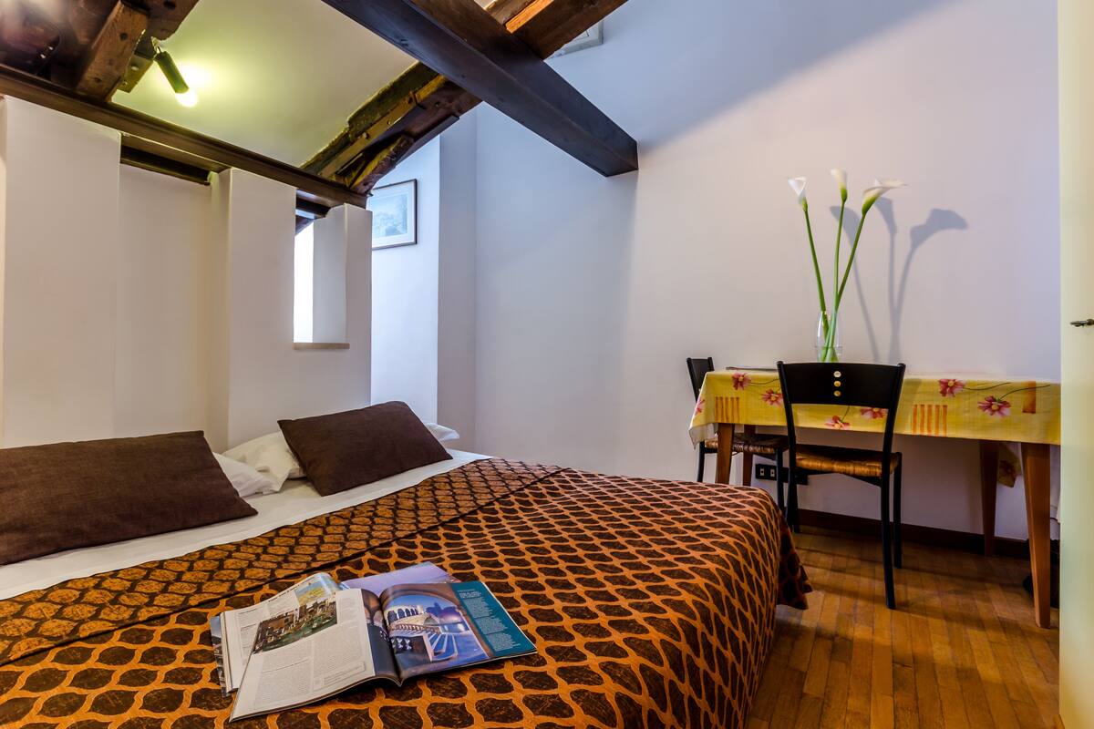 A bedroom with a patterned bedspread, a table with a floral tablecloth, and a vase with flowers.