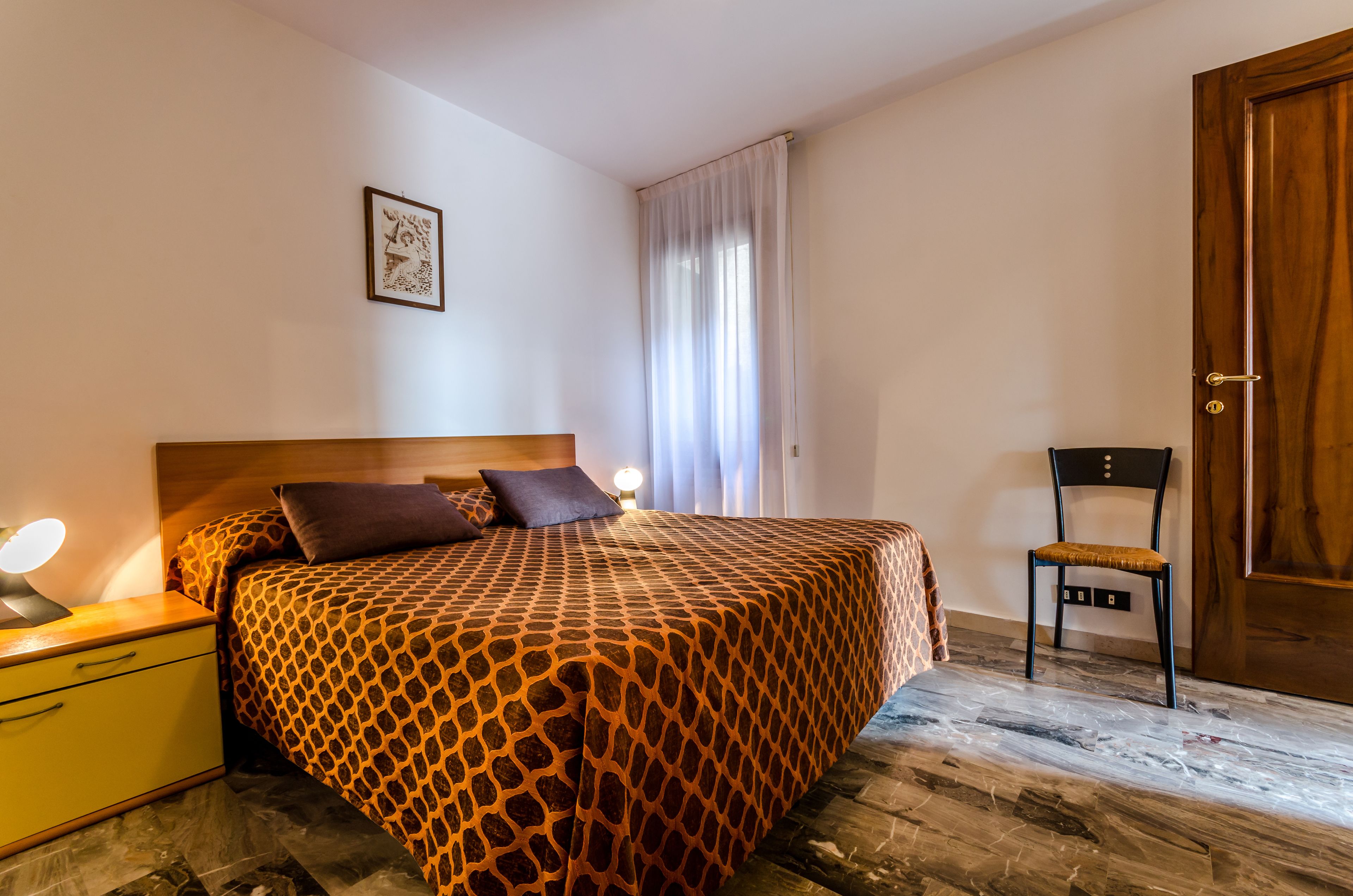 Una camera da letto con una coperta a fantasia, un comodino con una lampada, una sedia e una porta in legno.