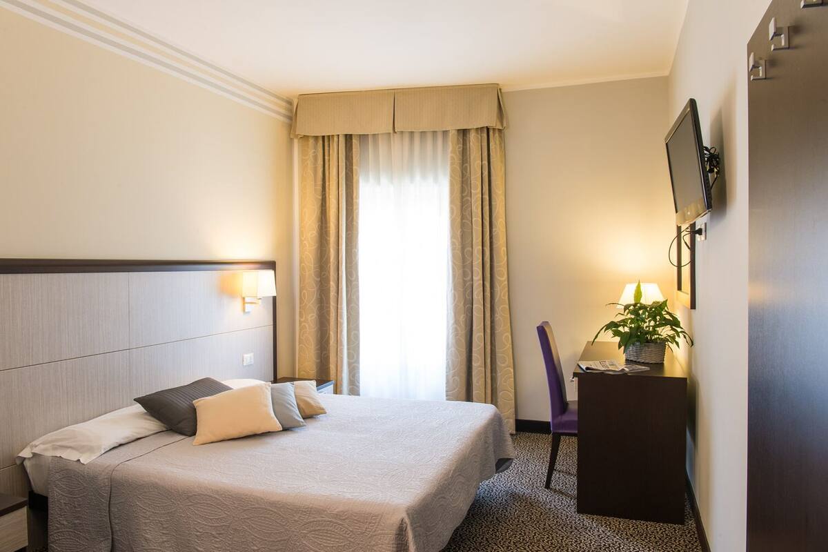 A hotel room with a bed, a desk with a lamp, a television, and a window with curtains.