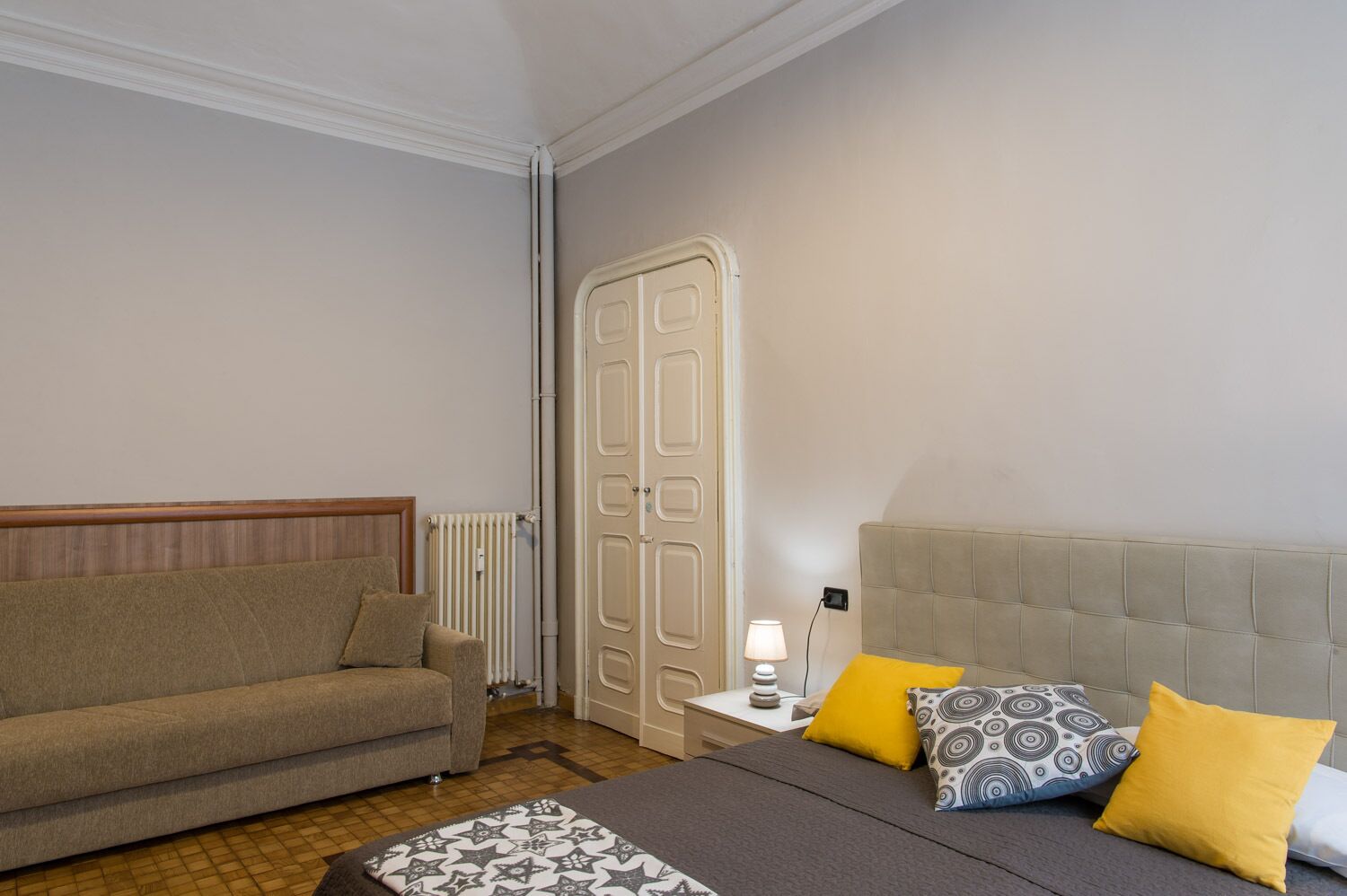 A hotel room with a bed, a sofa, a nightstand with a lamp, and a radiator.