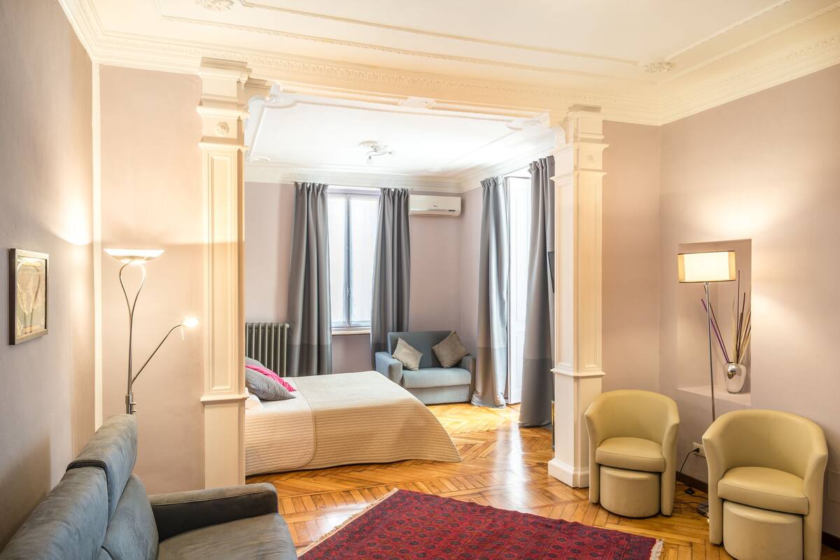 A hotel room with a bed, a sofa, two armchairs, a red rug, and a floor lamp.