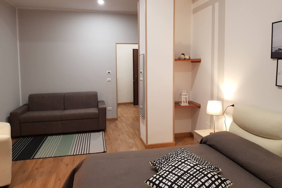 A modern bedroom with a bed, a sofa, a nightstand, a lamp, and a door leading to another room.