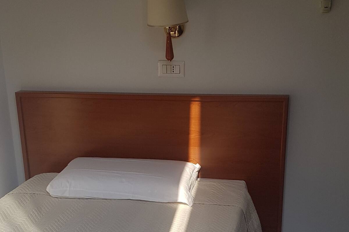 A neatly made bed with a wooden headboard, a wall-mounted lamp, and a thermostat on the wall.