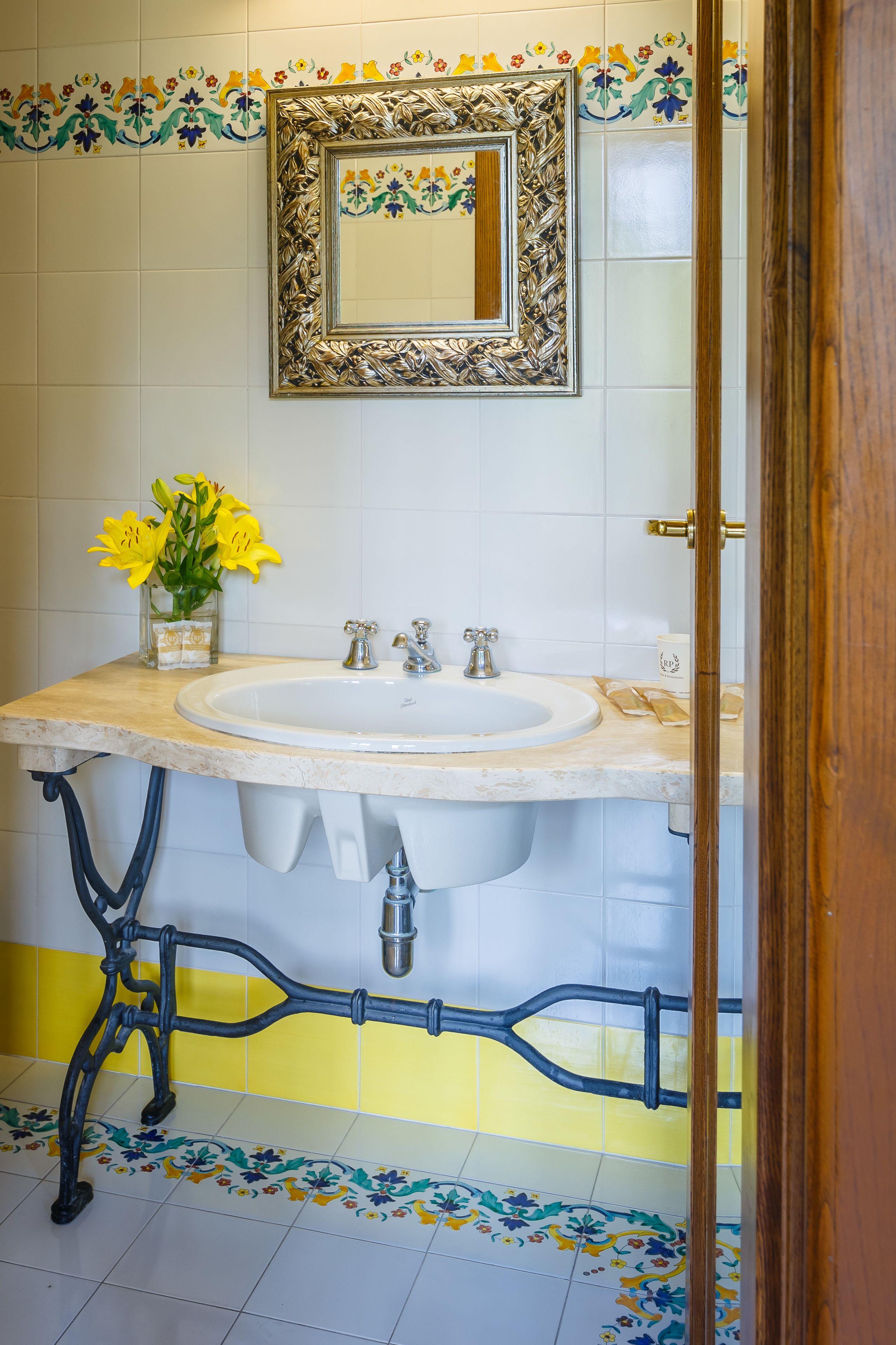 Een badkamer met een marmeren wastafel, een spiegel met een sierlijk lijstwerk en een vaas met gele bloemen.