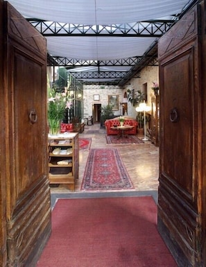 Un pasillo con alfombra roja que conduce a un área de estar con un sofá rojo y una mesa. Se observa un marco de puerta de madera a la izquierda y una pared de piedra a la derecha.