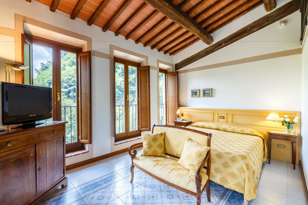 A bedroom with a bed, a sofa, a television on a wooden cabinet, and large windows.