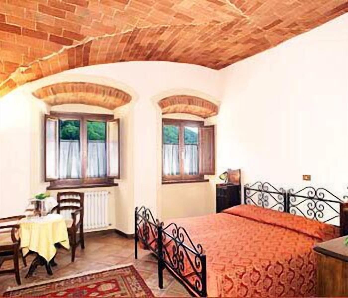 Un dormitorio con techo de ladrillo, dos ventanas con cortinas, una cama con estructura metálica, una mesita de noche y una silla.