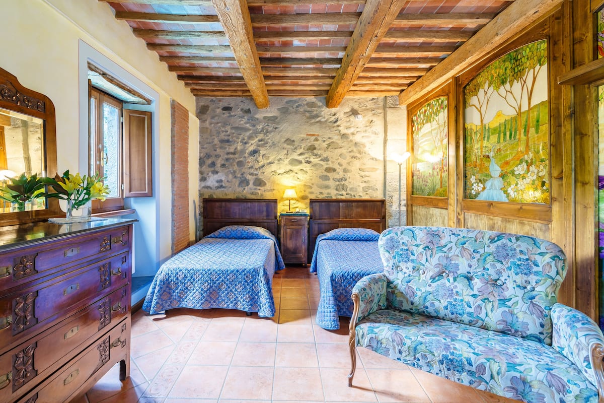 A rustic room with a stone wall, wooden beams, two beds with blue bedding, a wooden dresser, and a floral armchair.