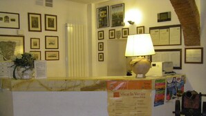 Zona de recepción de hotel con mostrador de mármol, cuadros en las paredes, una lámpara y un tablón de anuncios con horarios de eventos.