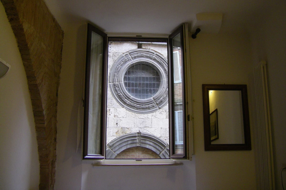 A room with a window overlooking a stone wall, a radiator, and a framed mirror on the wall.
