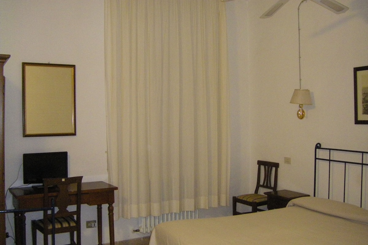 A hotel room with a bed, a desk with a chair, a television, a lamp, and a window with curtains.