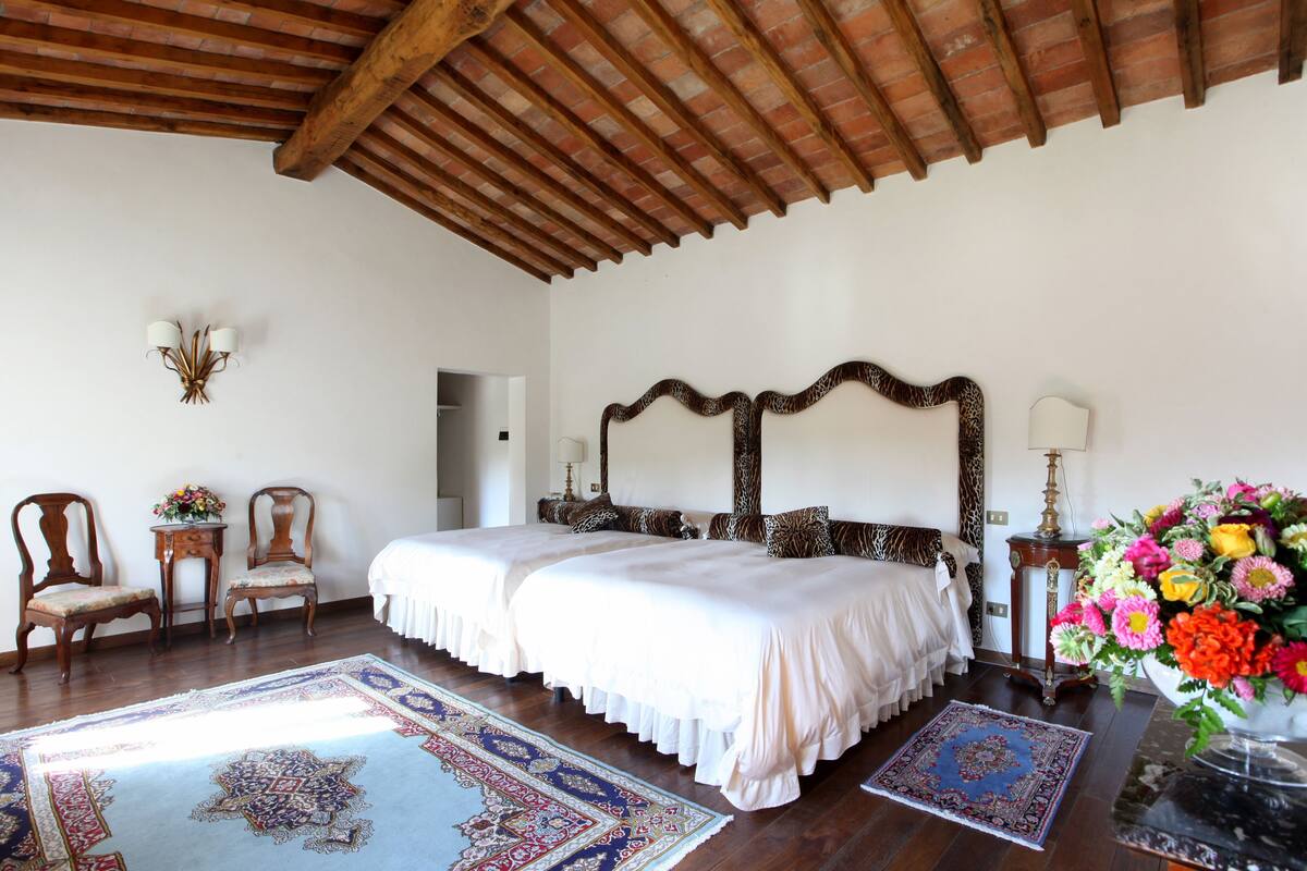 Un dormitorio con dos camas, sillas de madera, una mesa con flores y una alfombra grande.