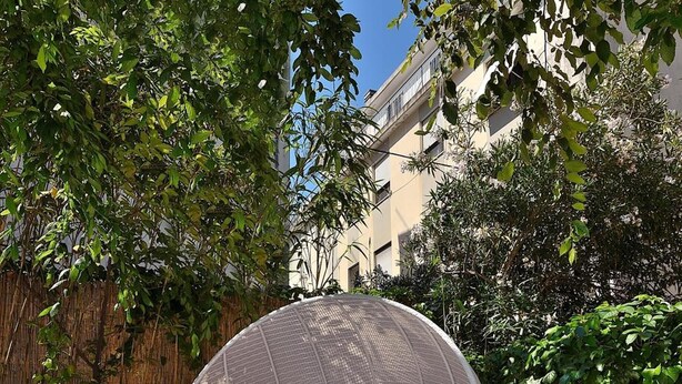 Ein runder, gewebter Sitzbereich im Freien mit orangefarbenen Kissen, einer großen Dekorationsvase und einem Bambuszaun in einem sonnigen Innenhof.