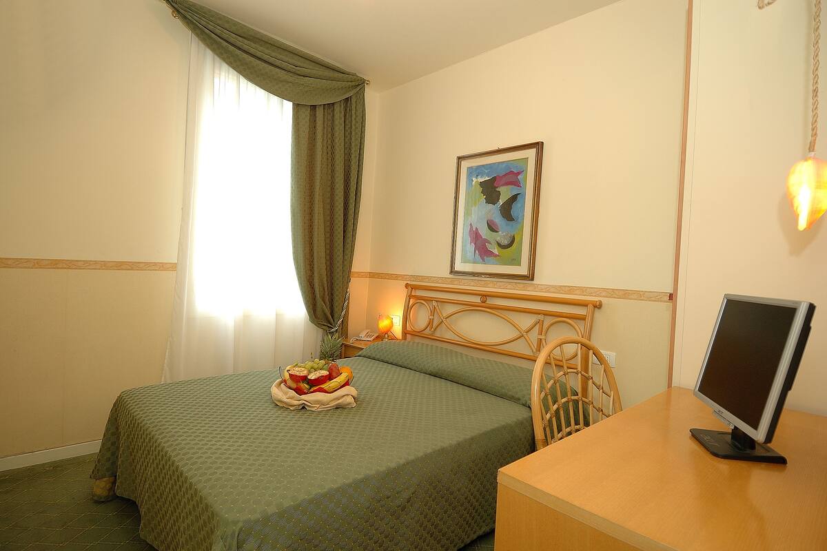 A hotel room with a bed, a desk with a computer monitor, a fruit bowl on the bed, and a painting on the wall.