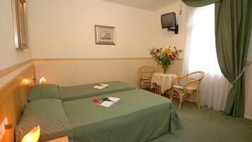 Una camera d'albergo con due letti, un tavolino con un vaso di fiori, due poltrone in vimini, un televisore a parete e una finestra con tende.