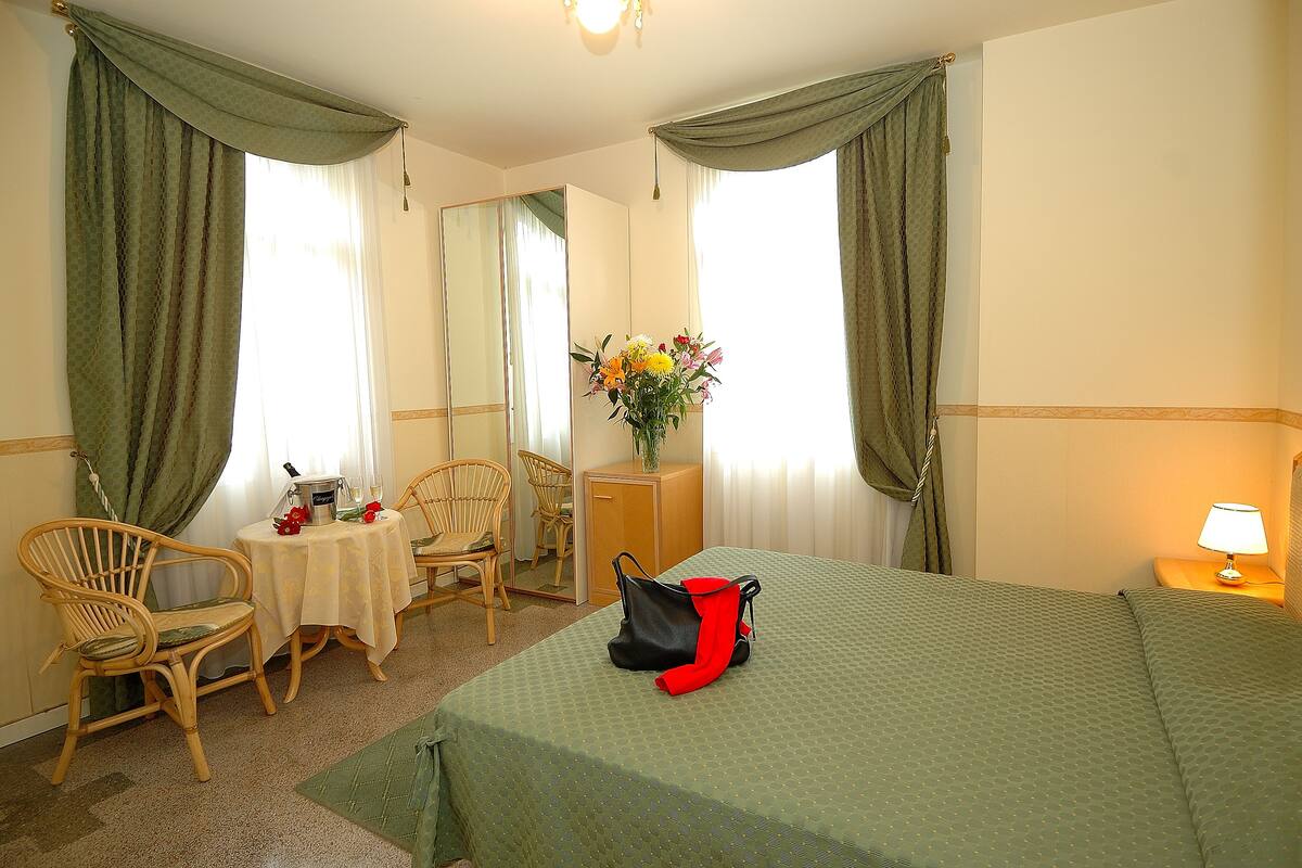 A hotel room with a bed, a desk with a chair, a mirror, a lamp, and a vase with flowers.