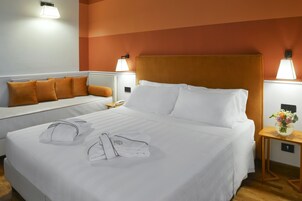 Habitación de hotel con una cama grande, una mesita de noche con lámpara, un pequeño sofá con cojines naranja y una lámpara de pared.