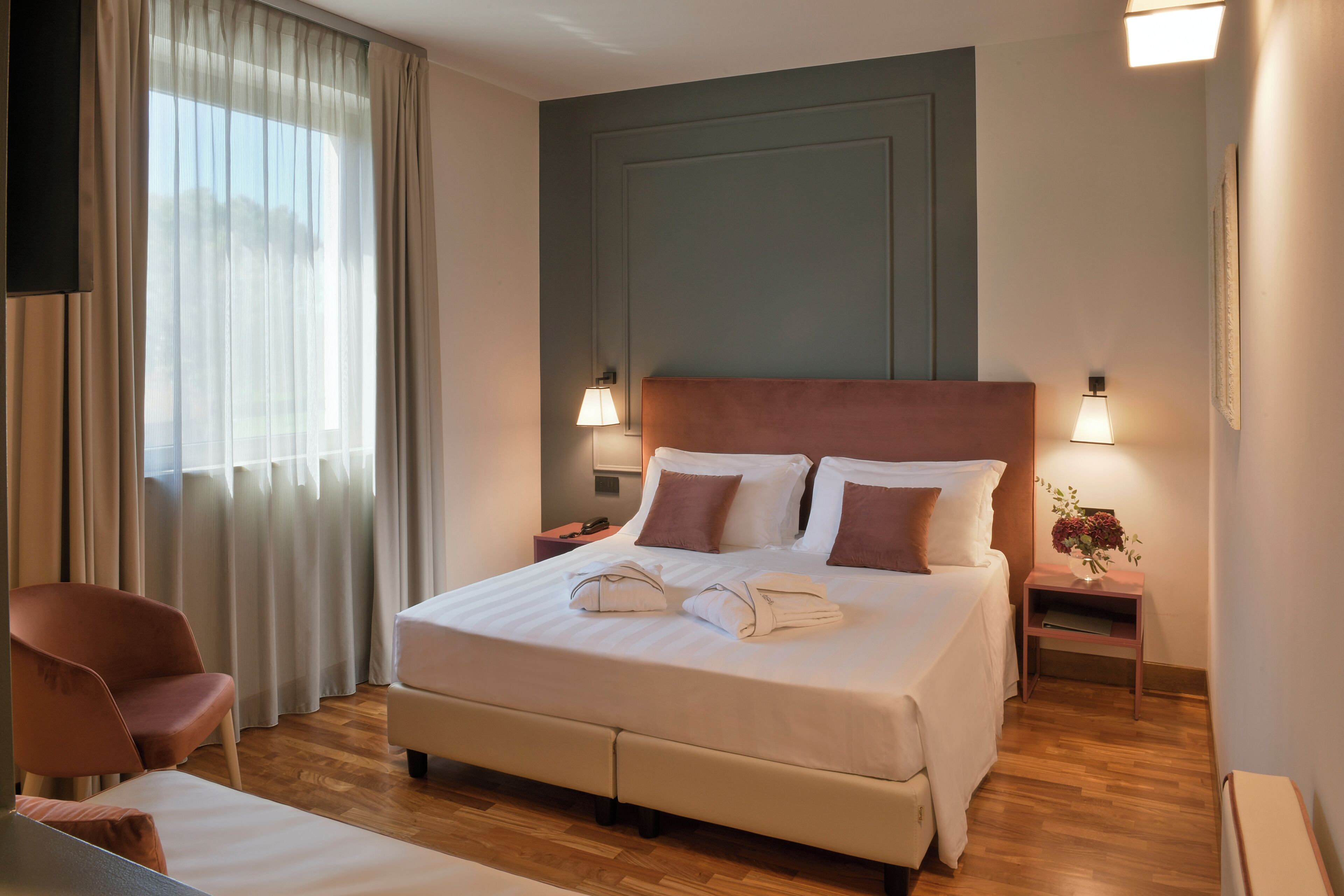Una camera d'albergo con un letto grande, due lampade ai comodini, una sedia, un comodino e una finestra con tende.