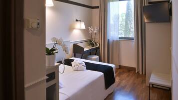 Ein Hotelzimmer mit einem Bett, einem Schreibtisch, einem Stuhl, einem kleinen Tisch, einer Topfpflanze, einer Lampe und einem Fernseher.