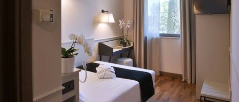 Habitación de hotel con cama, escritorio, silla, mesita, planta en maceta, lámpara y televisor.