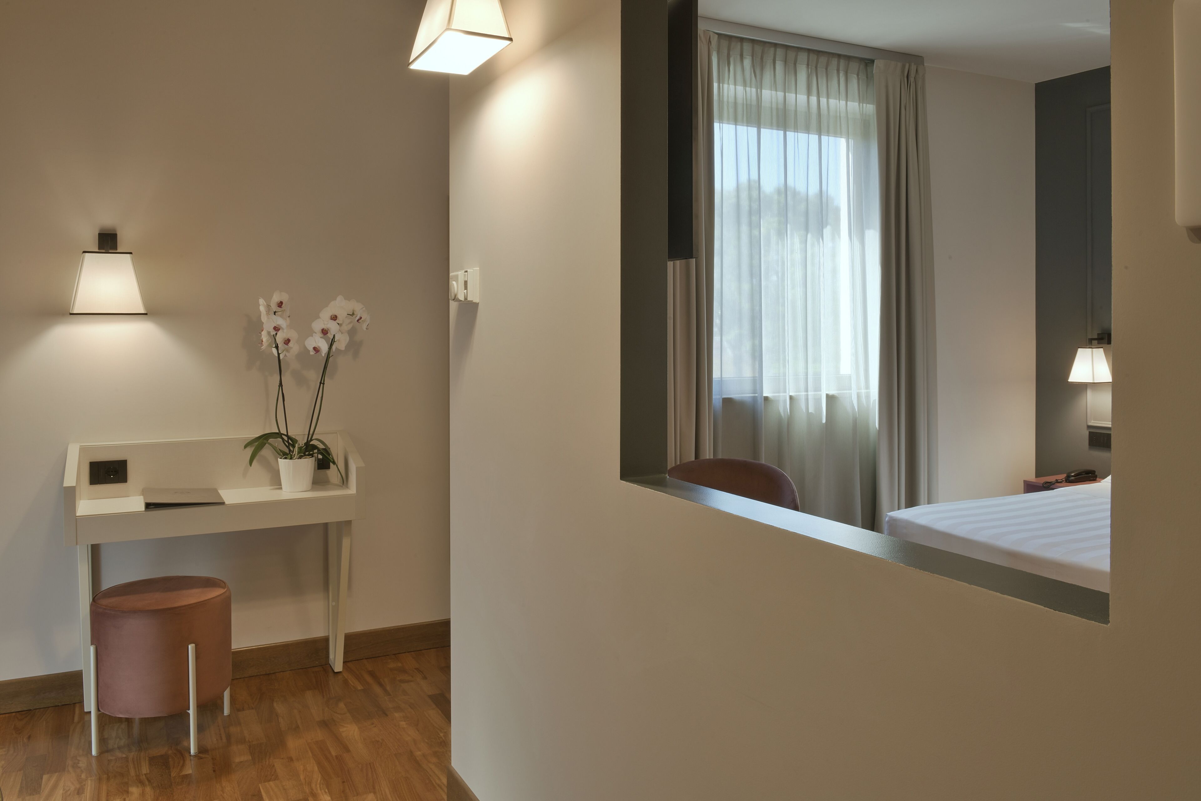 A modern hotel room with a white desk, a small pink stool, a potted orchid, a window with curtains, and a mirror.