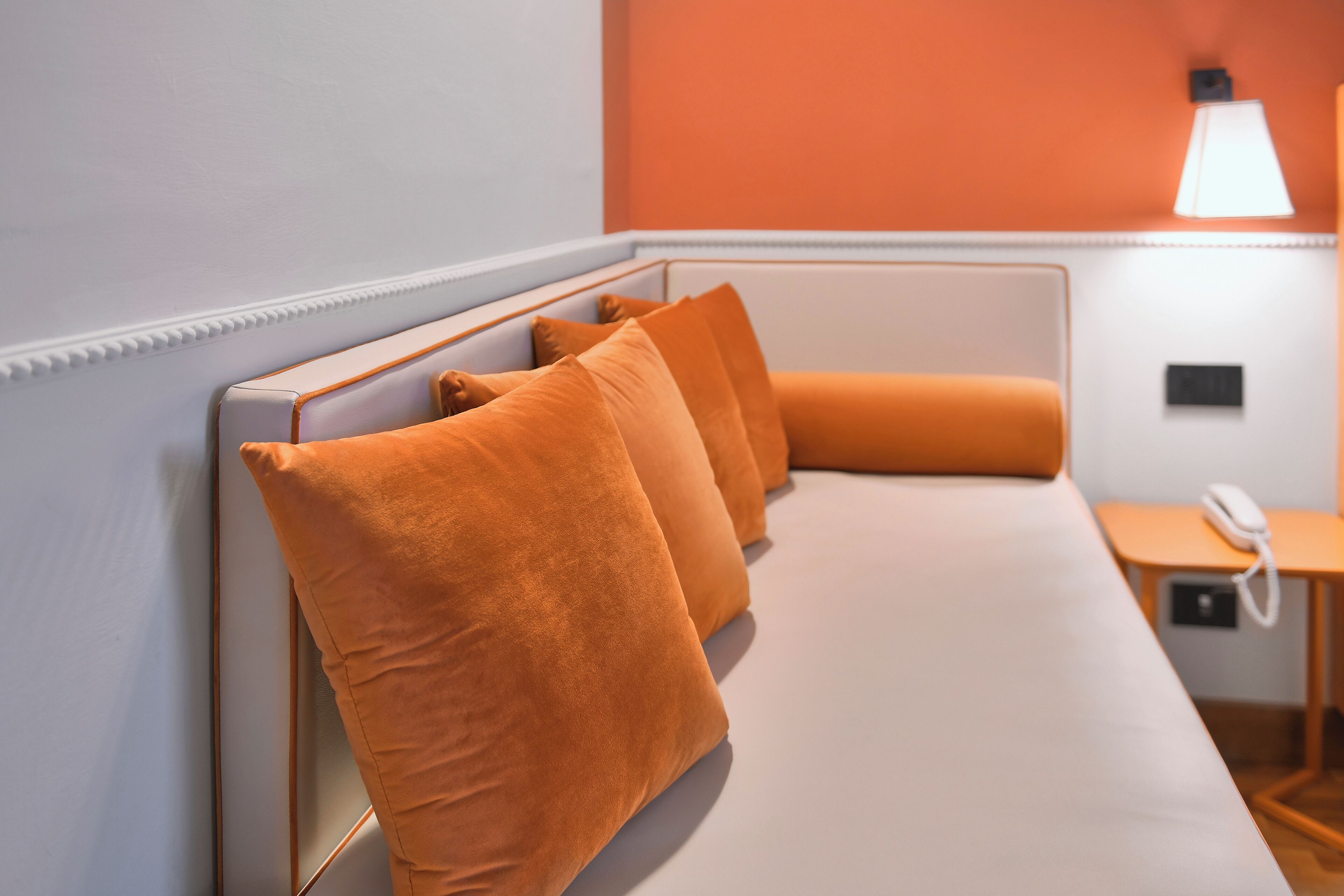 Ett hotellrum med en vit säng, orange kuddar, ett sängbord med en lampa och en telefon.