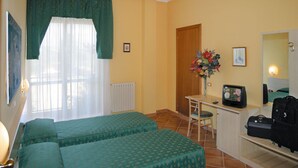 A room with two beds, a desk with a computer, a chair, a radiator, and a window with curtains.