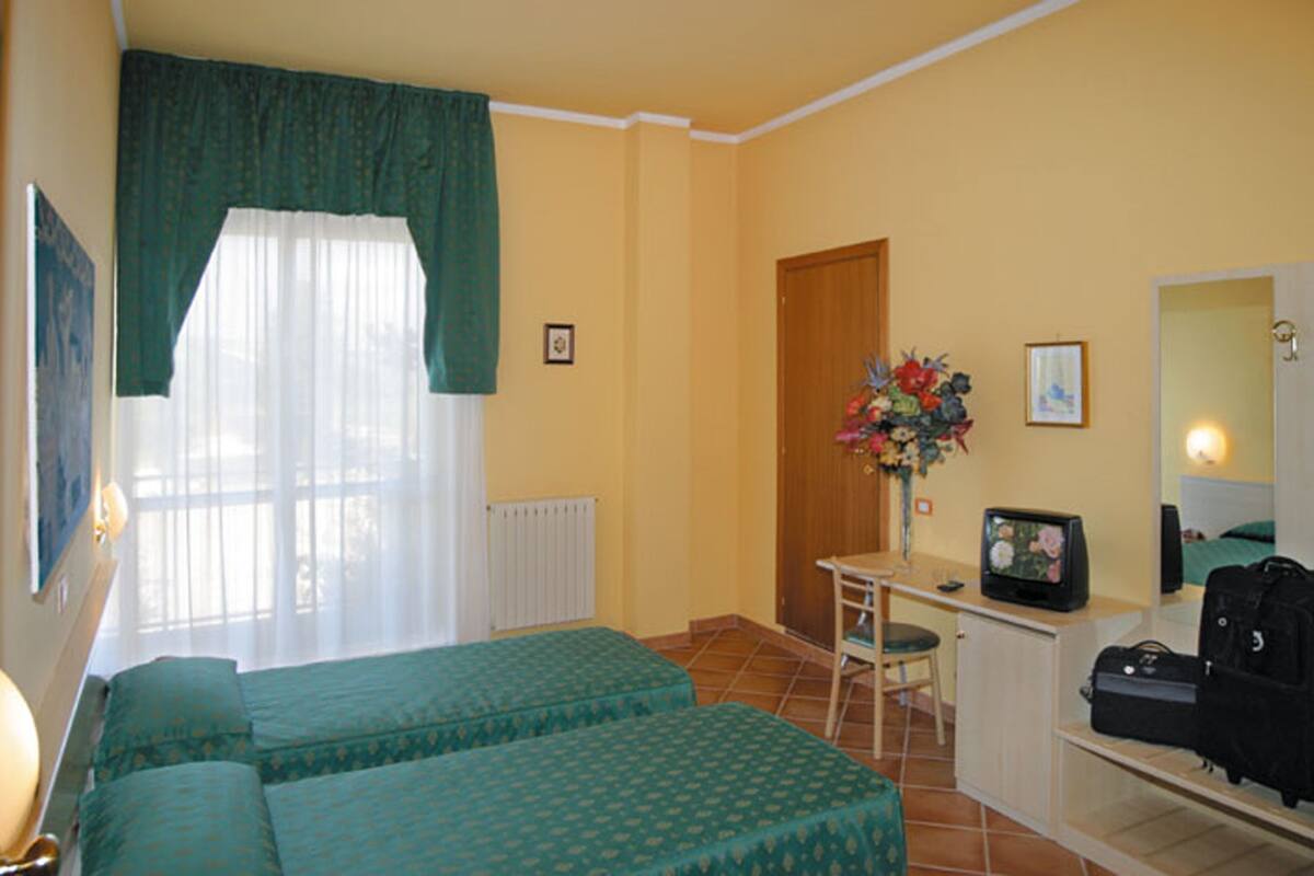 A room with two beds, a desk with a computer, a chair, a radiator, and a window with curtains.