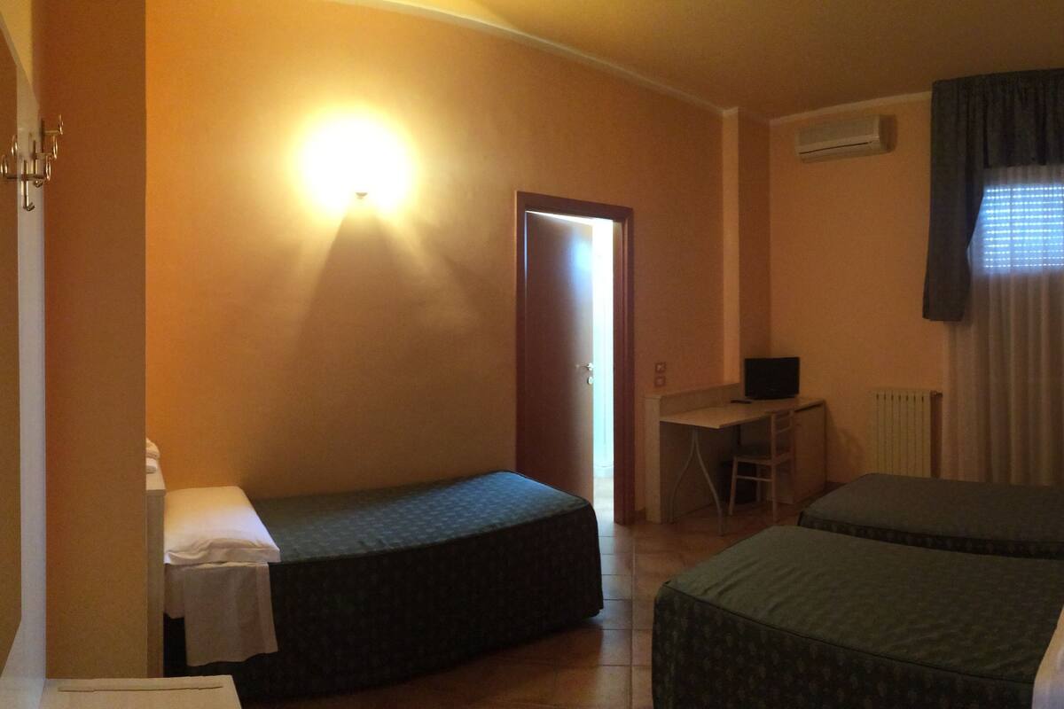 A small hotel room with two single beds, a desk with a chair, a computer, and a window with curtains.