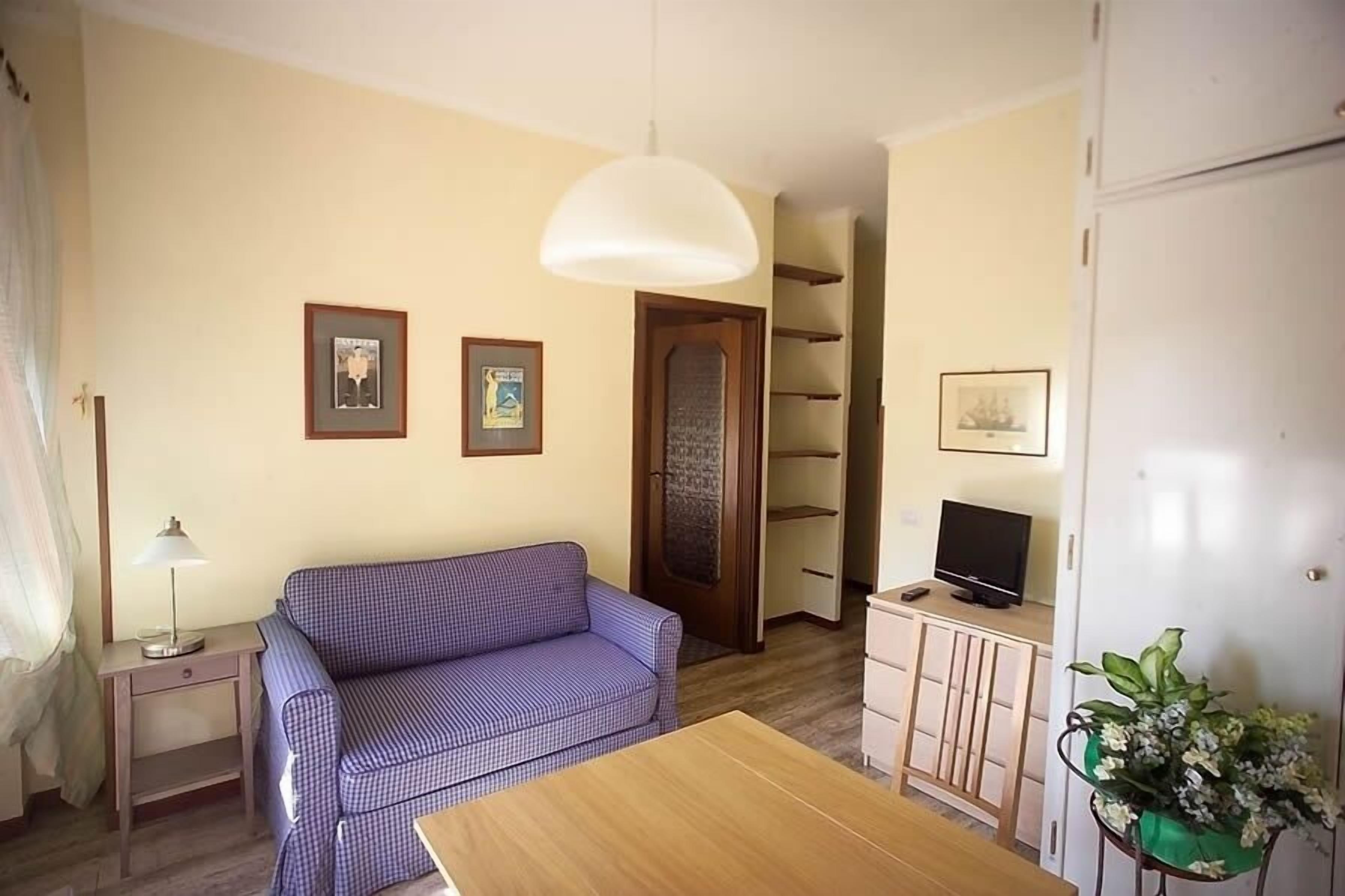 Una habitación con un sofá a cuadros, una mesa de madera, un televisor sobre un mueble y una puerta que da a un pasillo.