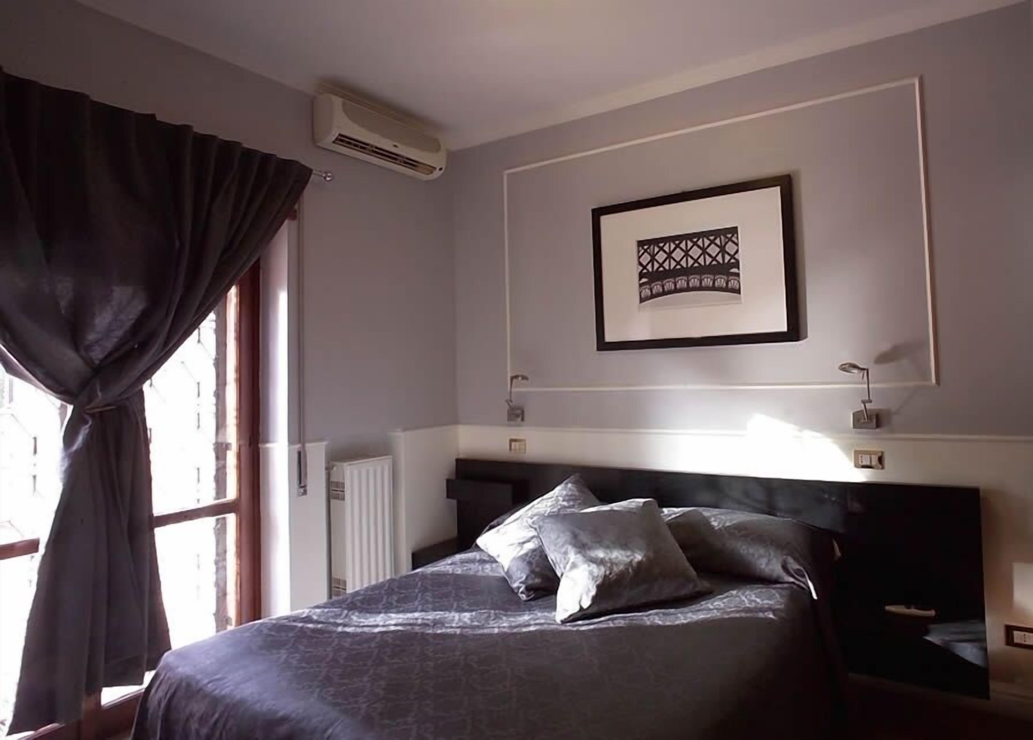 A bedroom with a bed, a window with curtains, an air conditioning unit, a framed picture, and bedside lamps.