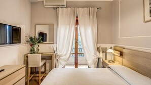 A hotel room with a bed, a desk with a chair, a TV, a bedside table with a lamp, and a window with curtains.