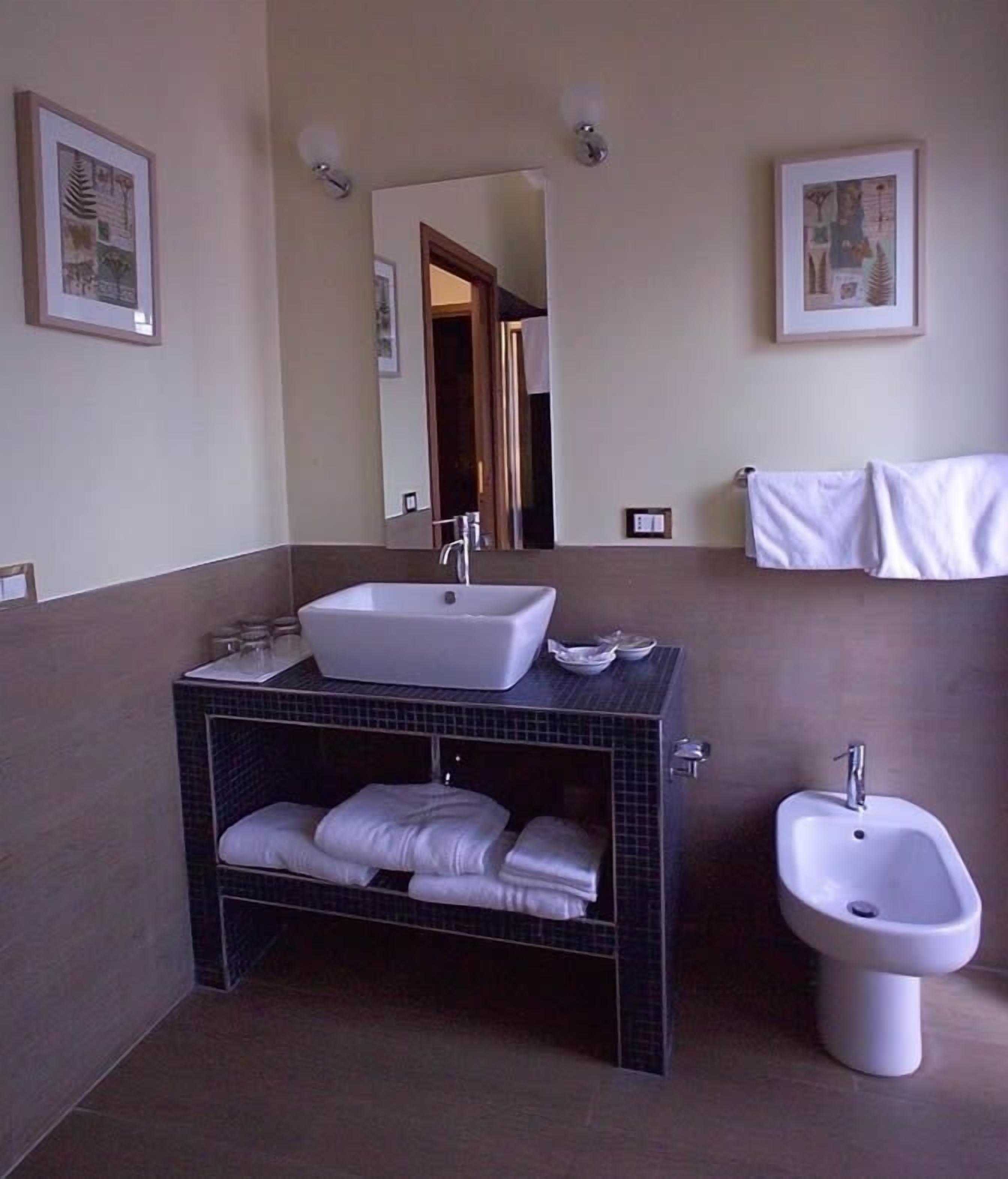 Ein Badezimmer mit einem großen Waschbecken, einem Bidet und einem Spiegel. Handtücher hängen an der Wand und ein Regal zeigt gefaltete Handtücher.