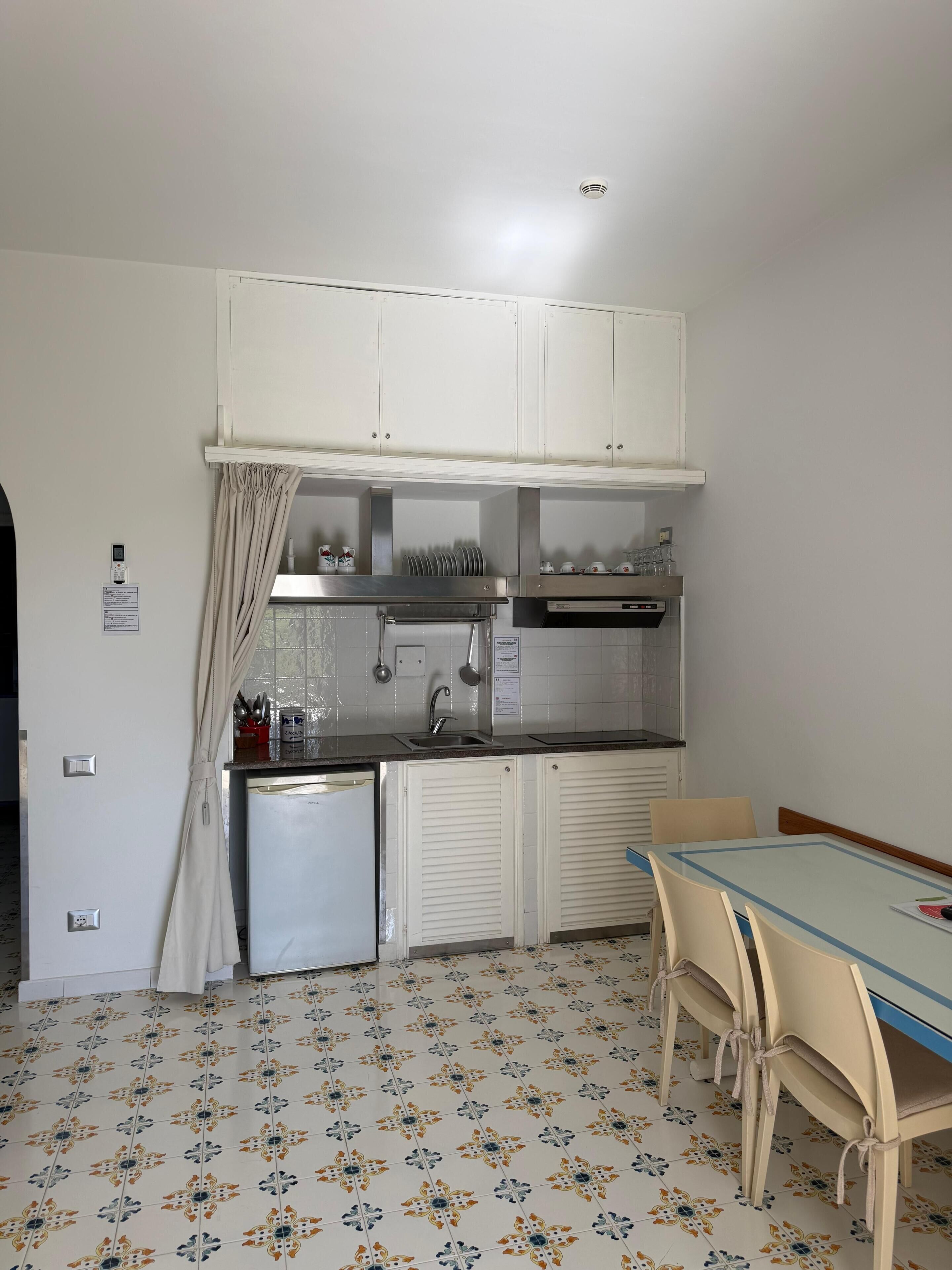 A compact kitchen with white cabinets, a small refrigerator, a sink, and a dining table with chairs.